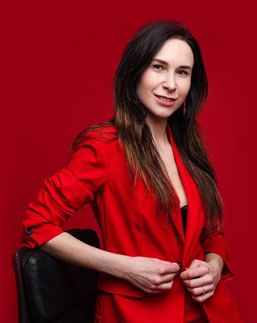 Portrait of caucasian female with long dark straight hair in red office suit, black shoes sits on black chair